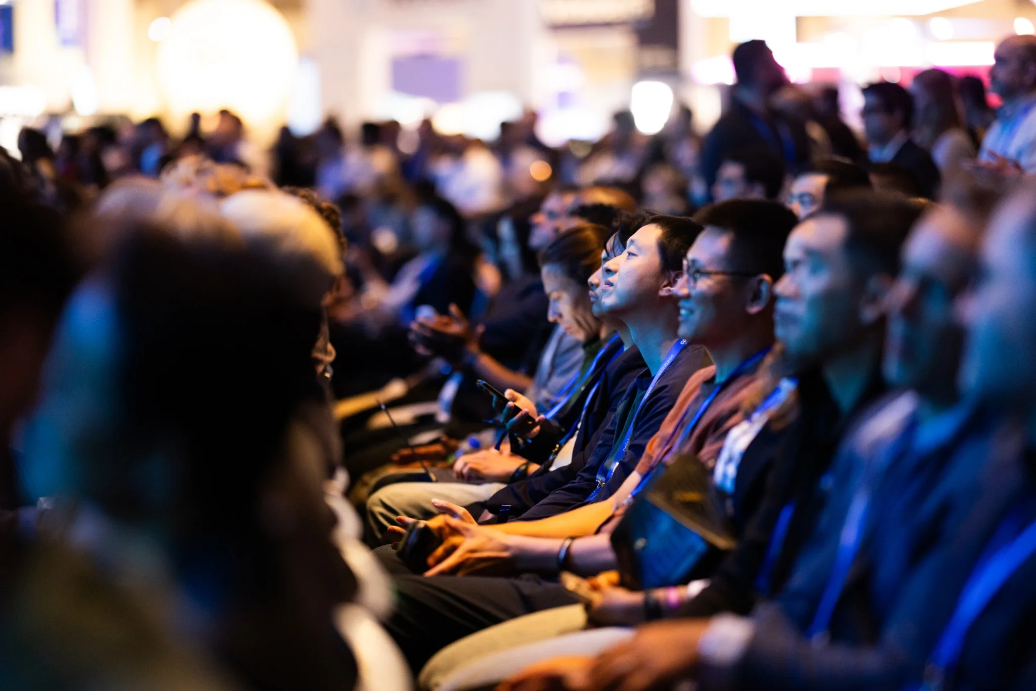 Rows of seated attendees wearing lanyards watching a presentation under warm blue event lighting