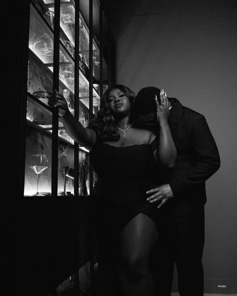 Couple beside a backlit glassware cabinet, woman in off-shoulder dress reaching for the frame