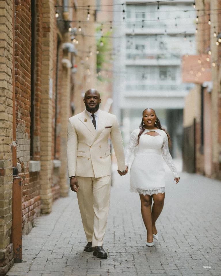 Couple in cream suit and short white dress strolling hand in hand down a brick alley
