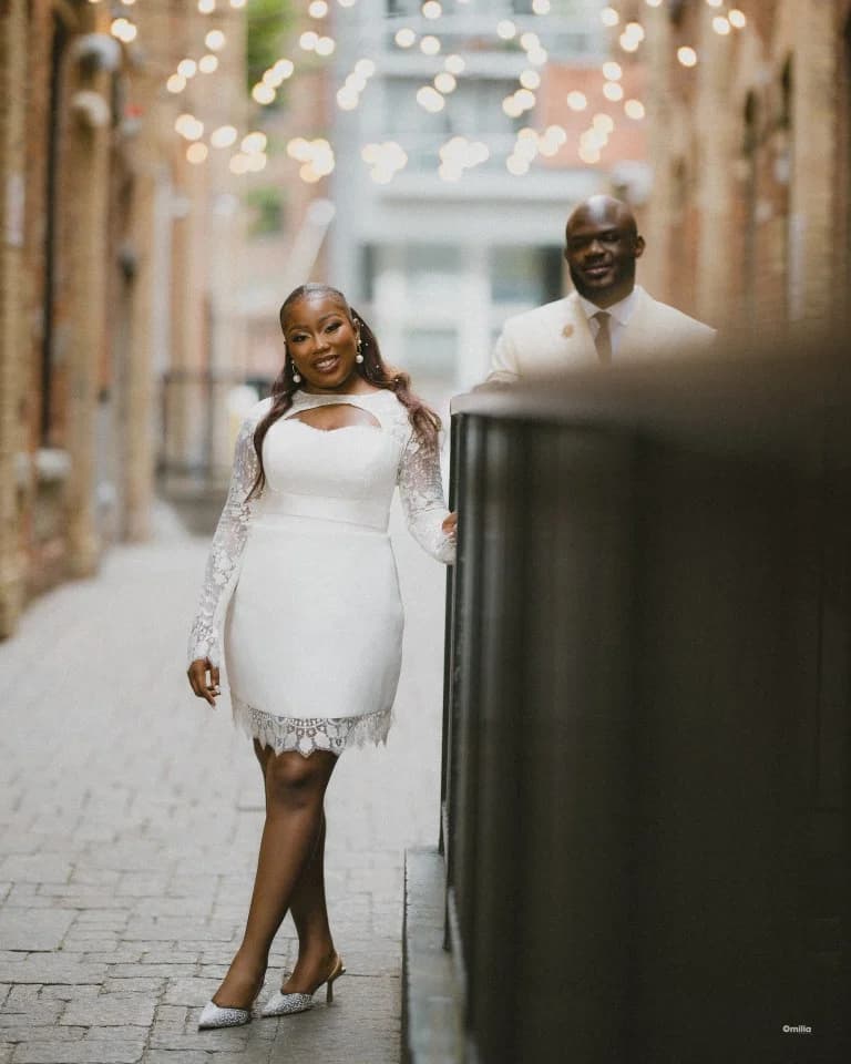 Woman in short lace dress and jeweled heels posing in cobbled alley, partner behind