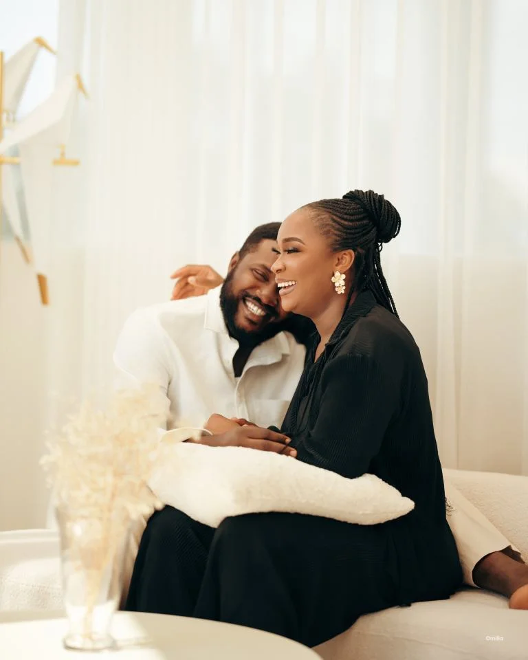 Laughing couple nestled cheek-to-cheek on cream sofa beside sheer sunlit curtains
