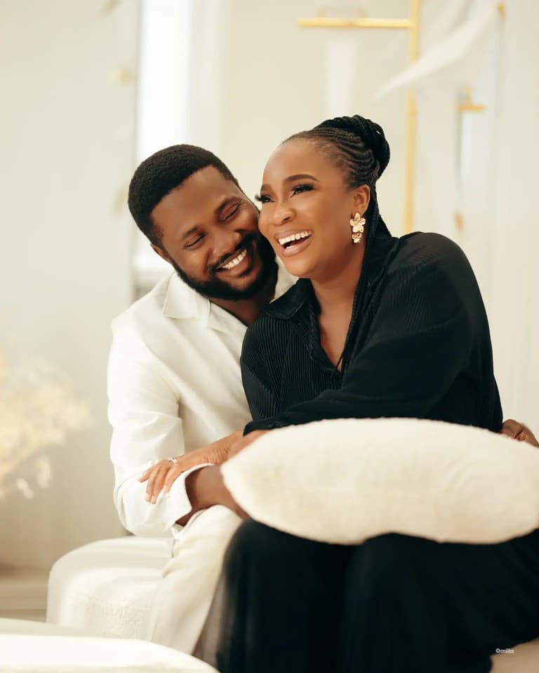 Couple laughing while seated indoors, he in white shirt embracing her in black pleated outfit