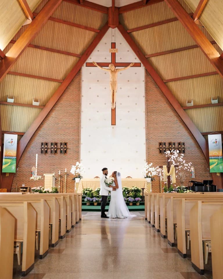Newlyweds embracing at flower-filled altar beneath crucifix in soaring A-frame sanctuary