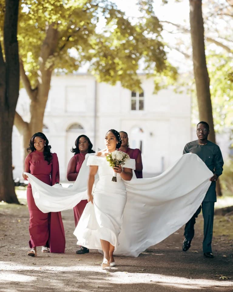 Bride walking a sunlit path while attendants in burgundy dresses carry her flowing train