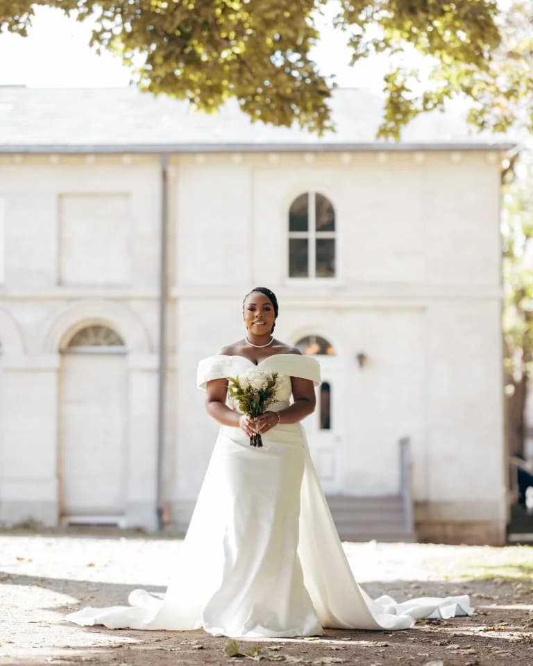 Bride in off-shoulder gown holding a white bouquet before a pale stone building