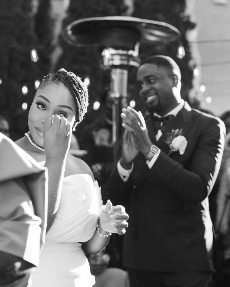 Bride wiping away a tear while the smiling groom applauds behind her