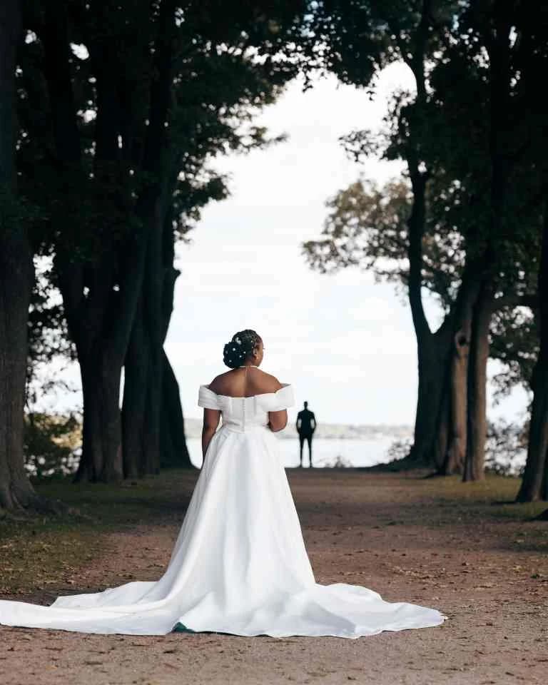 Bride seen from behind on a tree-lined path, groom waiting in the distance