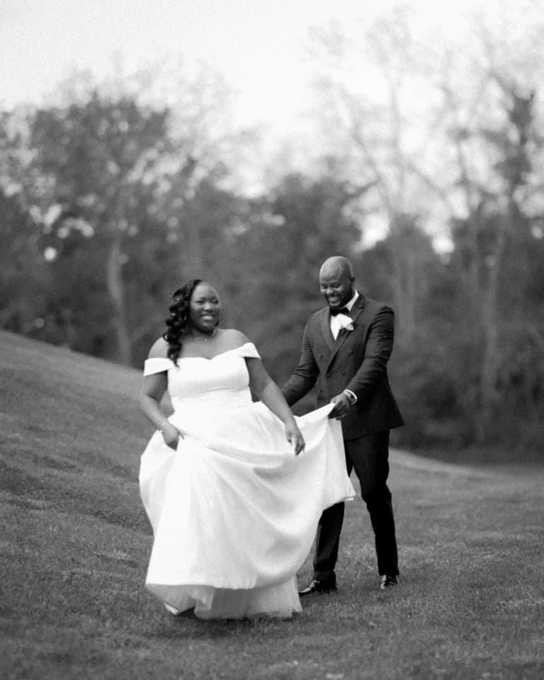 Groom lifting the bride's gown train as they walk across a grassy slope