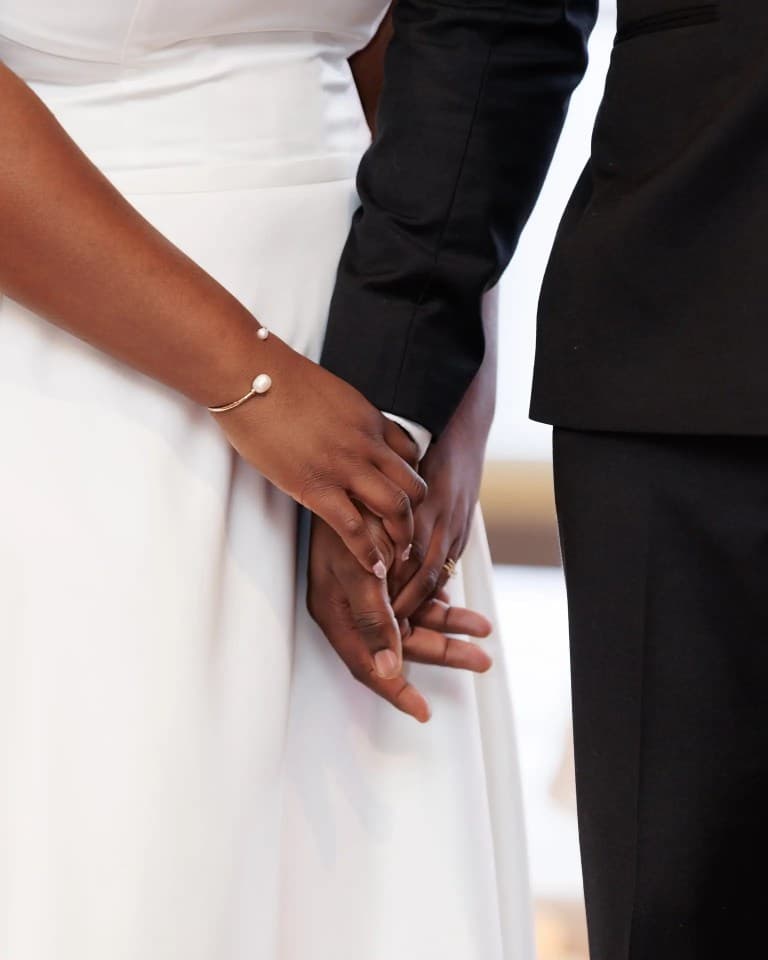 Bride and groom's clasped hands in close-up, pearl bracelet against the white gown