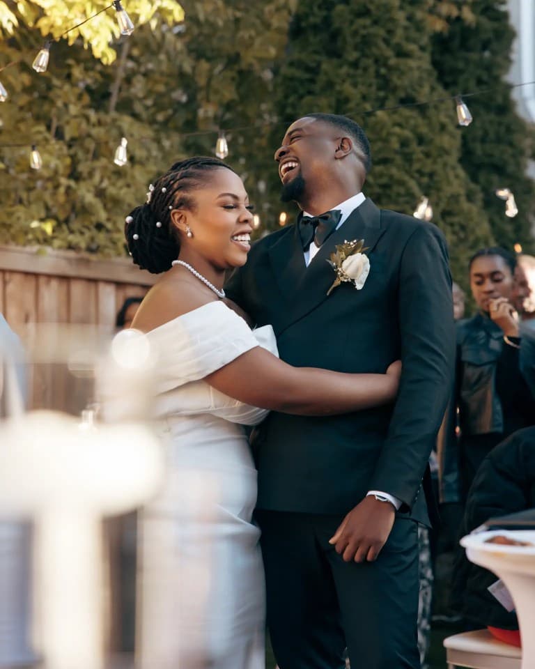 Groom laughing as the bride embraces him beneath string lights at outdoor reception