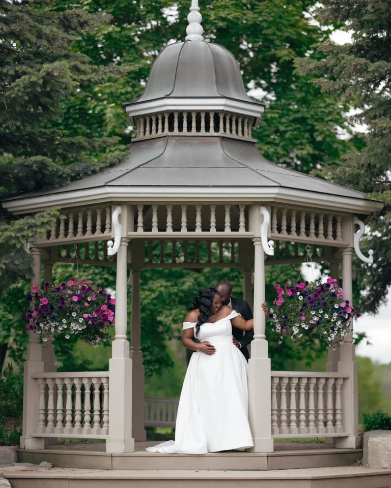 Groom embracing bride from behind inside gazebo hung with petunia baskets