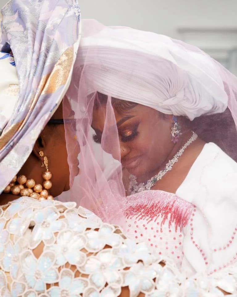 Bride in white gele sharing tearful embrace with woman in floral headwrap