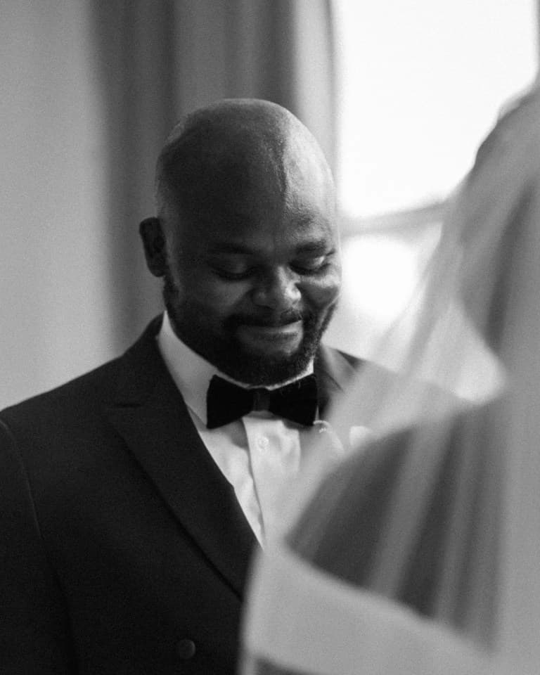 Groom in tuxedo and velvet bow tie smiling softly beside window light