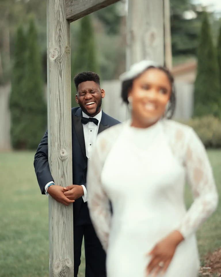 Groom laughing beside a wooden pergola post as the bride stands softly blurred in front