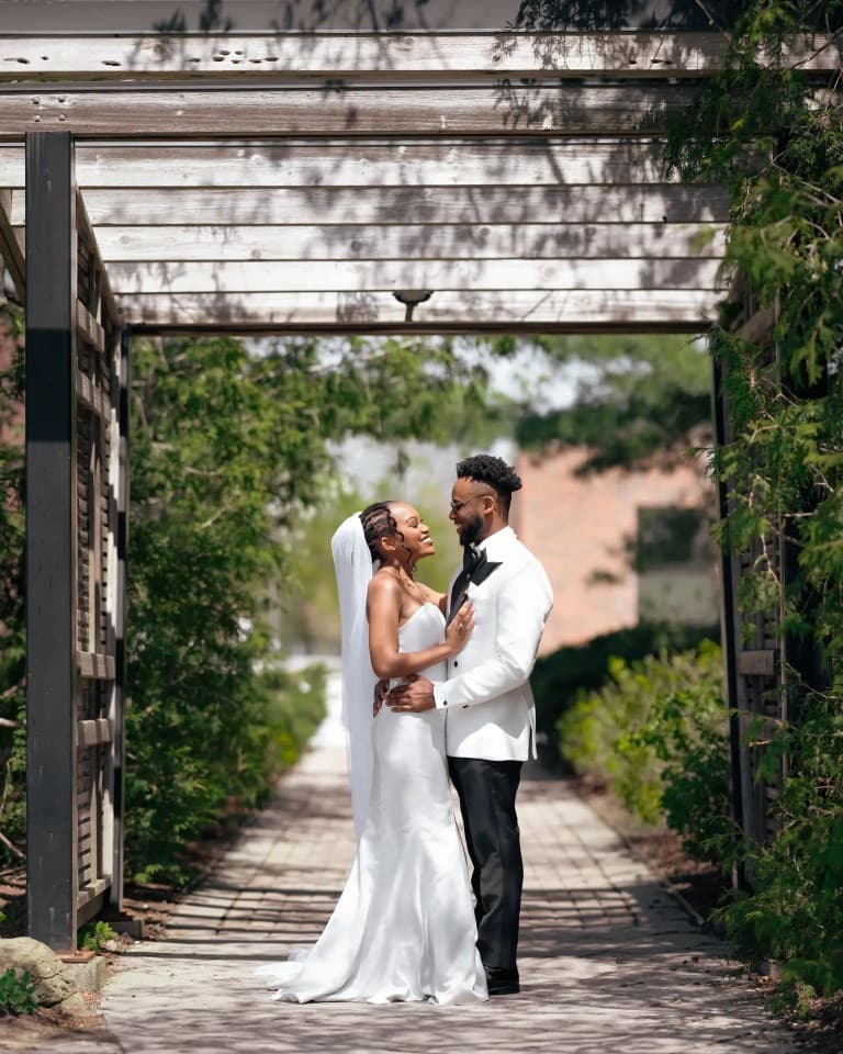 Bride and groom in white dinner jacket embracing beneath rustic wooden pergola