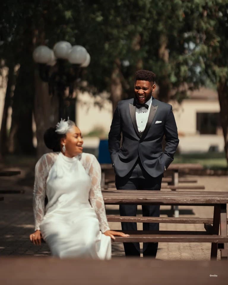 Bride seated at a wooden picnic table while the groom laughs behind her