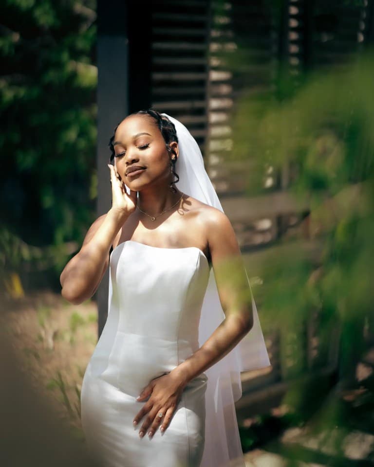 Bride in strapless satin gown and veil standing with eyes closed in dappled sunlight