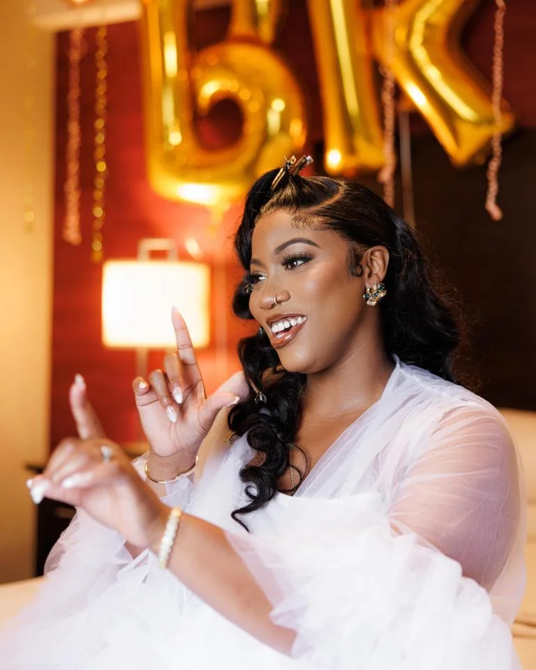 Smiling bride in sheer tulle robe gesturing playfully before gold letter balloons