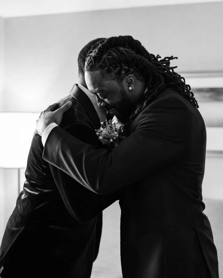 Two men in tuxedos sharing tight emotional embrace with rose boutonniere visible