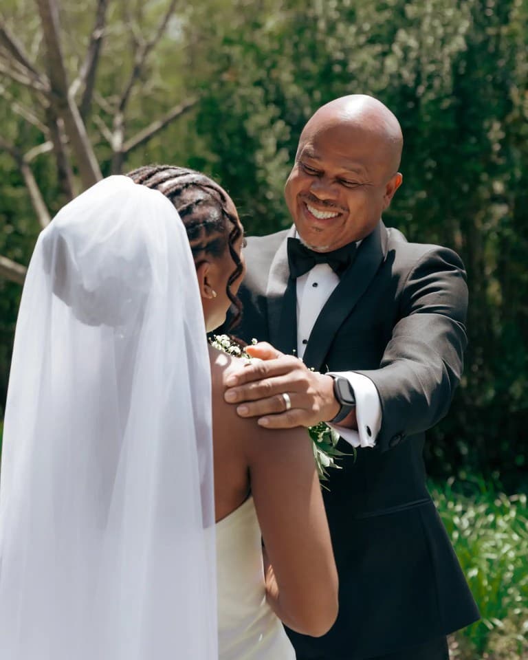 Bride in long veil embraced by smiling man in black tuxedo amid sunlit greenery