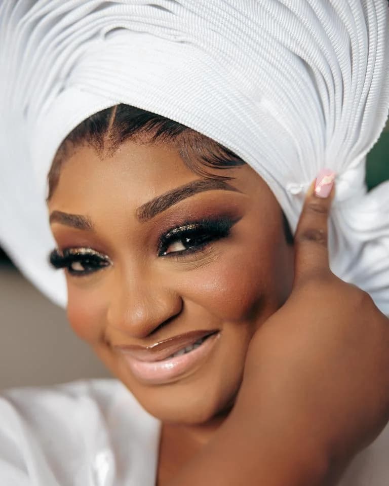 Bride in pleated white gele smiling with glamorous eye makeup, hand at cheek