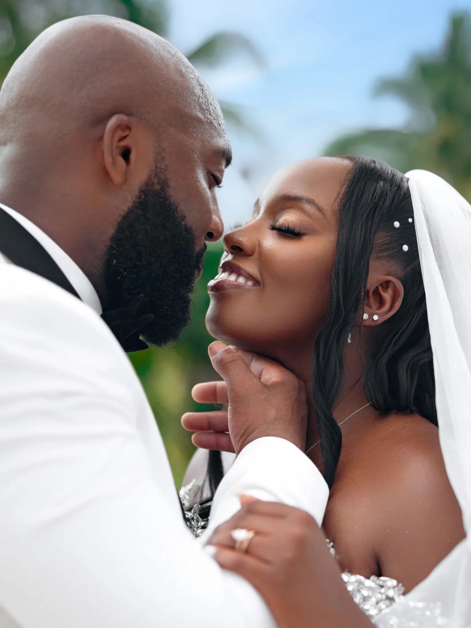 Groom in white tuxedo jacket cradling the smiling bride's chin under palm trees