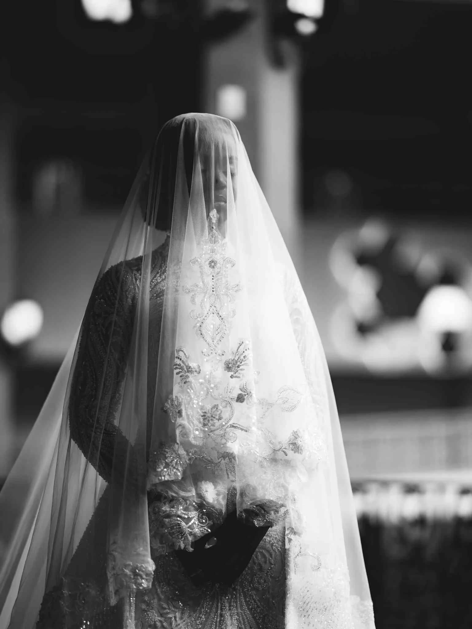 Bride standing with an embroidered veil draped over her face, holding a clutch