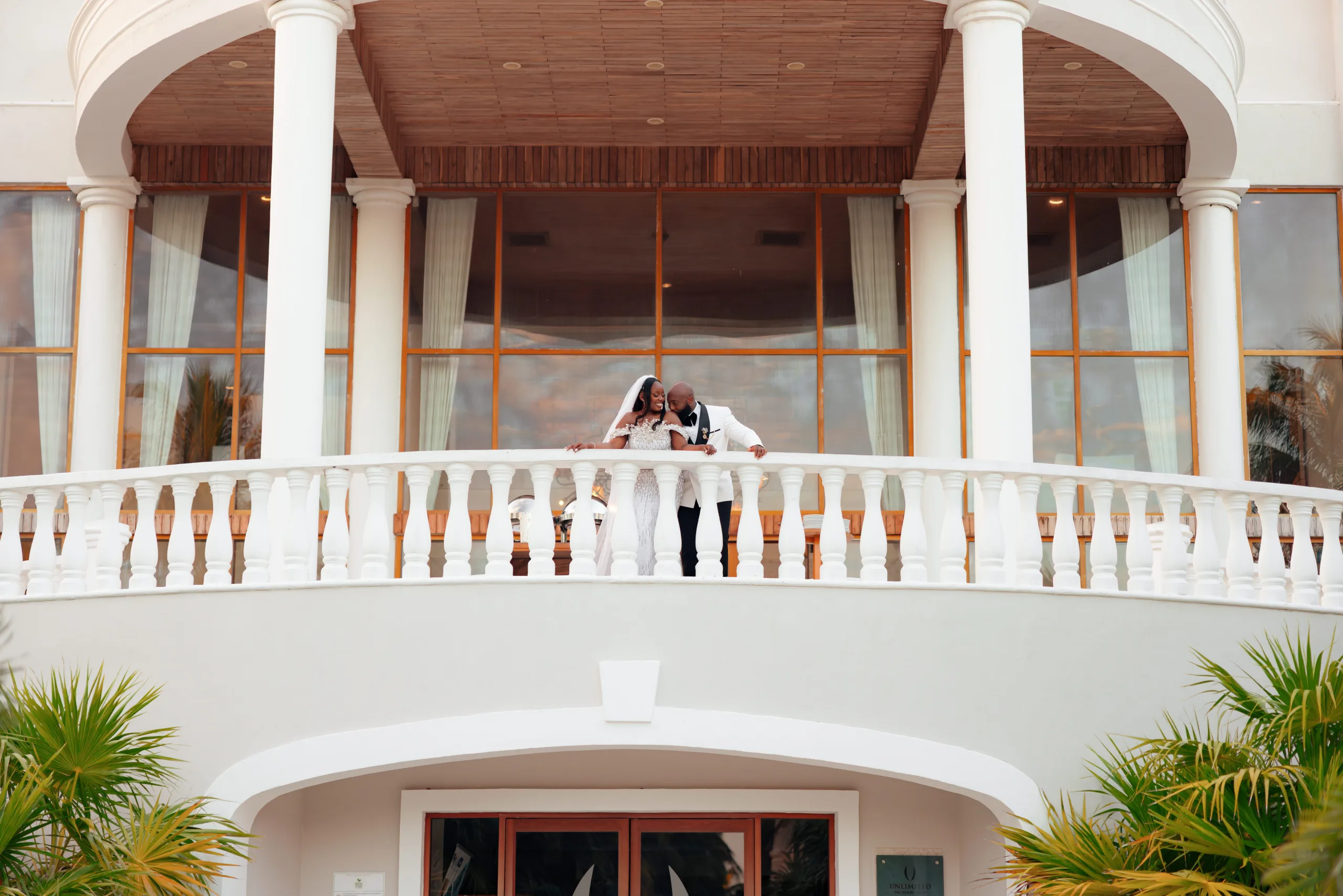 Bride and groom in white leaning close on curved balcony balustrade of grand columned building