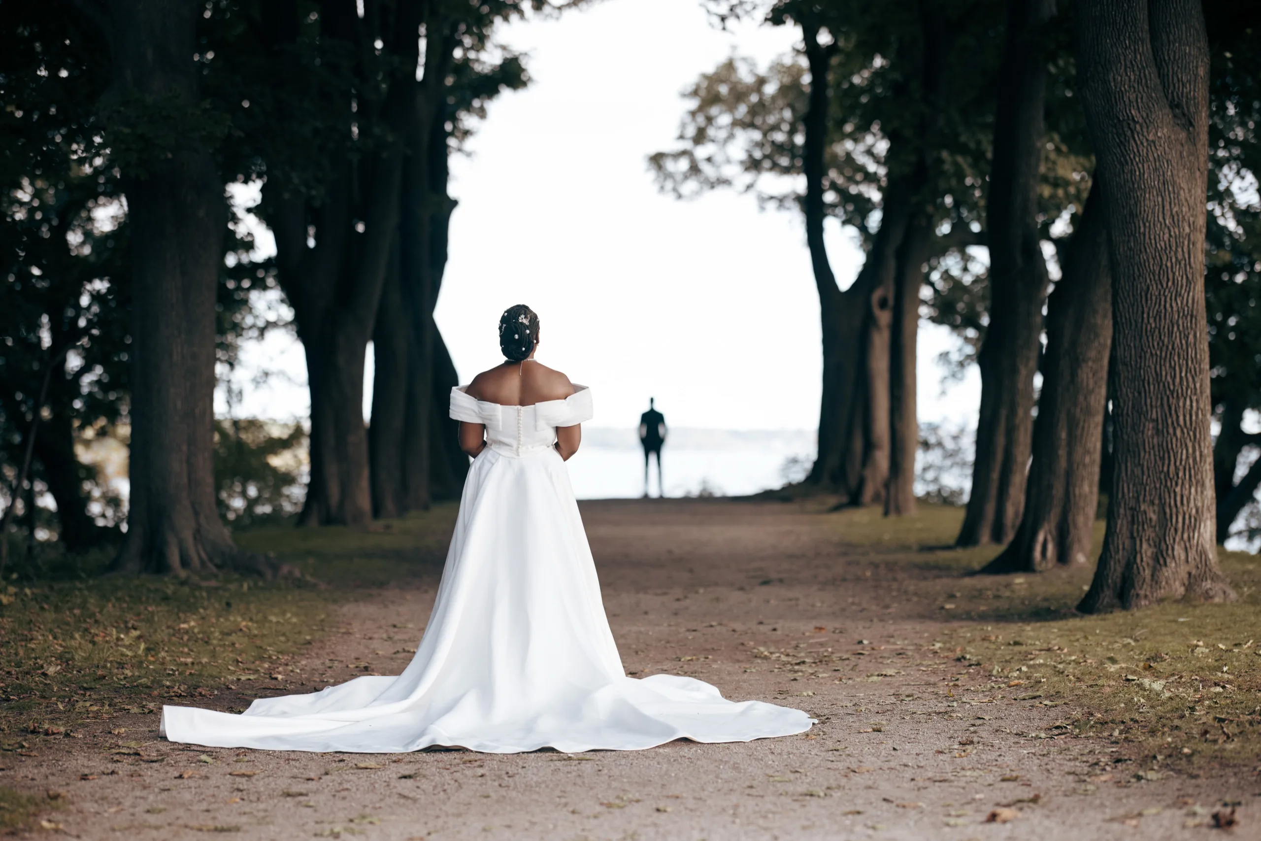 Bride facing down a tree-lined path toward the groom waiting in the distance for their first look