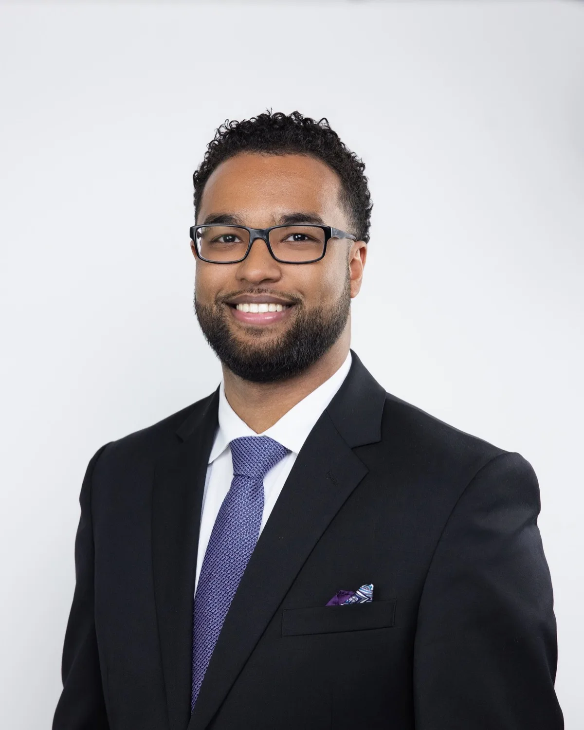 Smiling bearded man in black suit, purple tie, and glasses against light studio backdrop