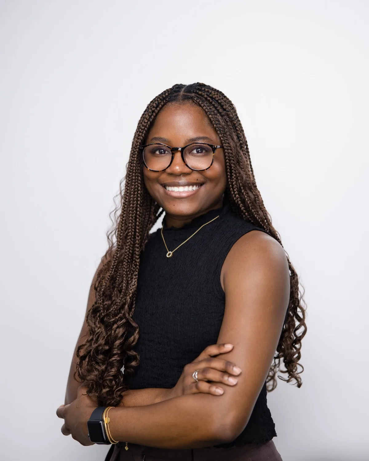 Woman with long braids and glasses in black sleeveless top, arms crossed, against white studio backdrop