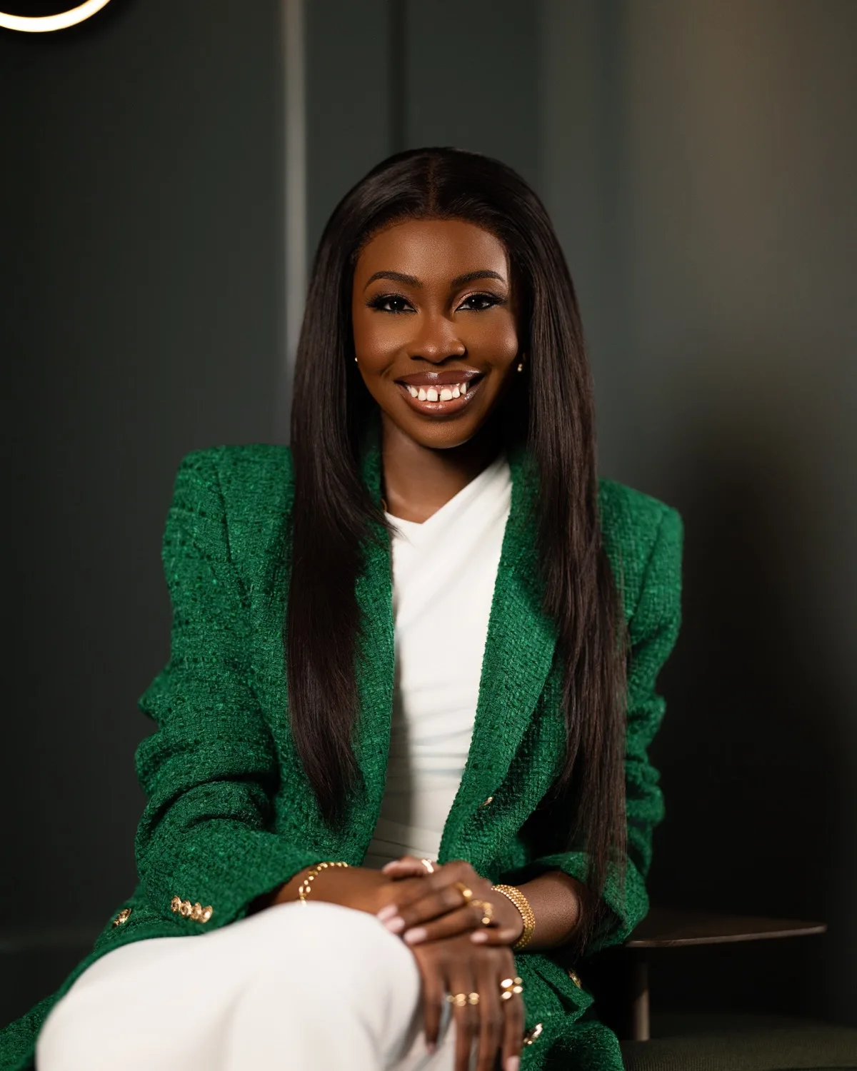 Smiling woman in green tweed blazer and white outfit seated against a dark green backdrop