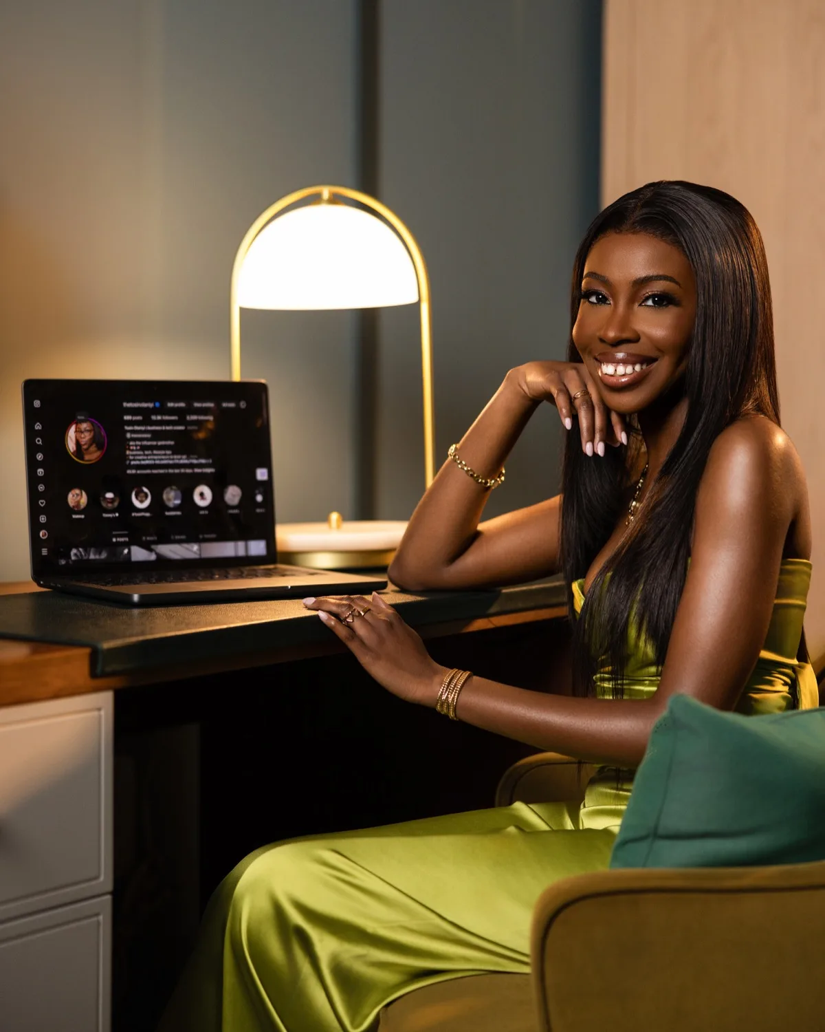 Smiling woman in green satin outfit seated at desk with laptop and glowing lamp, warm lighting