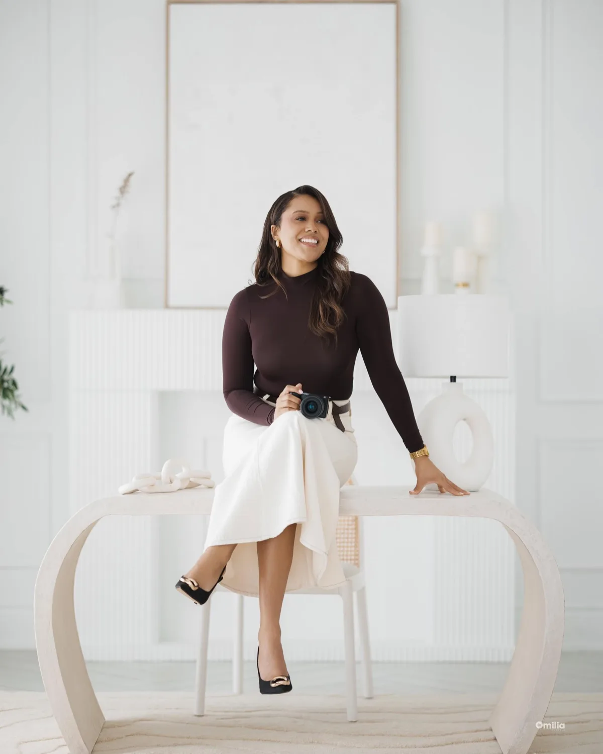 Woman in brown top and white skirt holding camera, seated on table in bright minimalist room