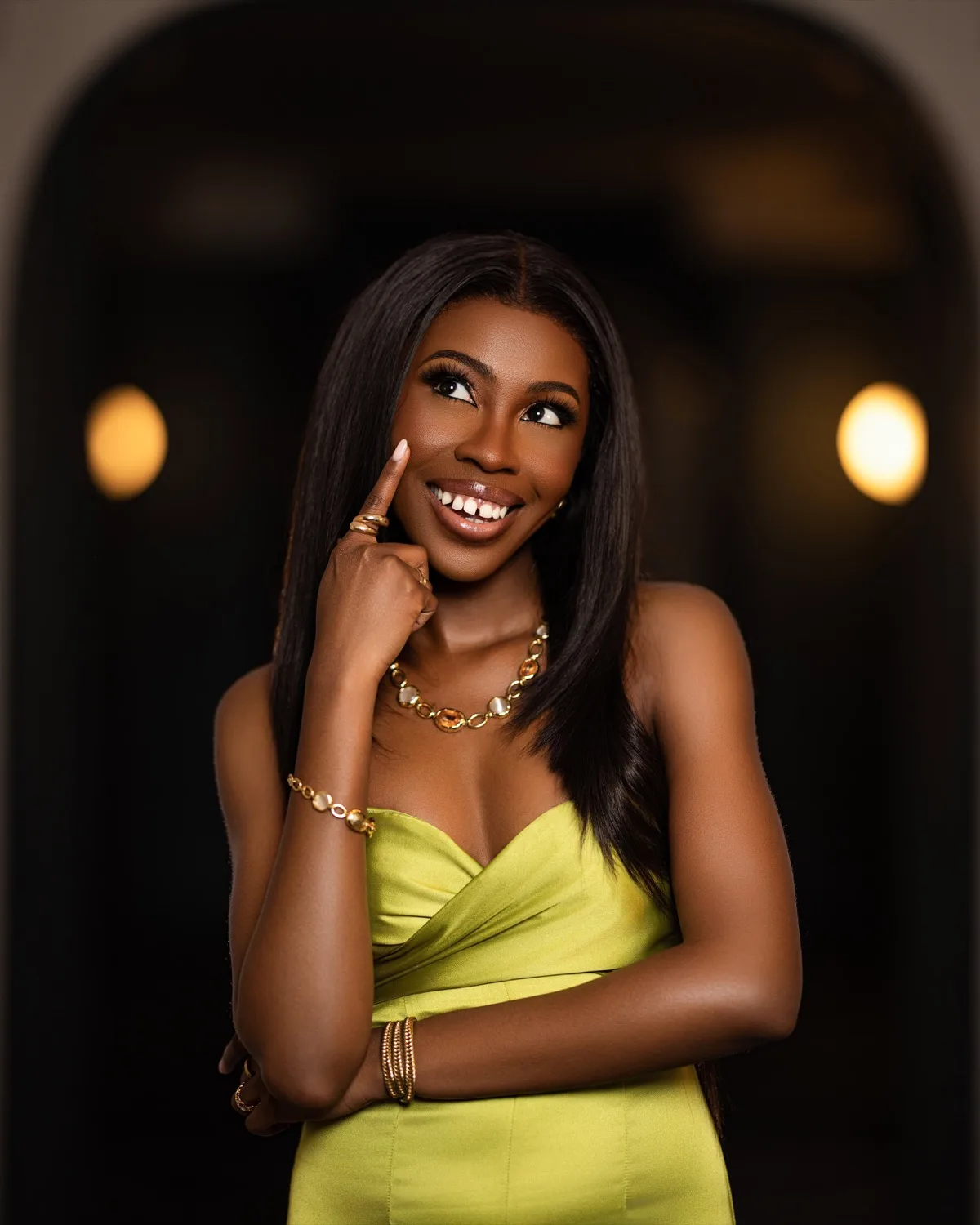 Smiling woman in lime satin strapless dress with gold jewelry, finger to cheek, warm bokeh background