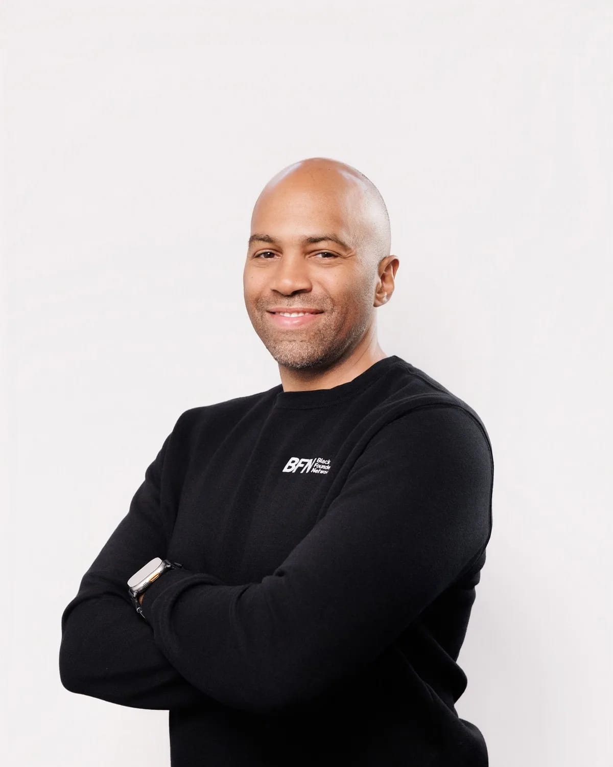 Smiling bald man with light stubble, arms crossed in black sweater against white studio backdrop