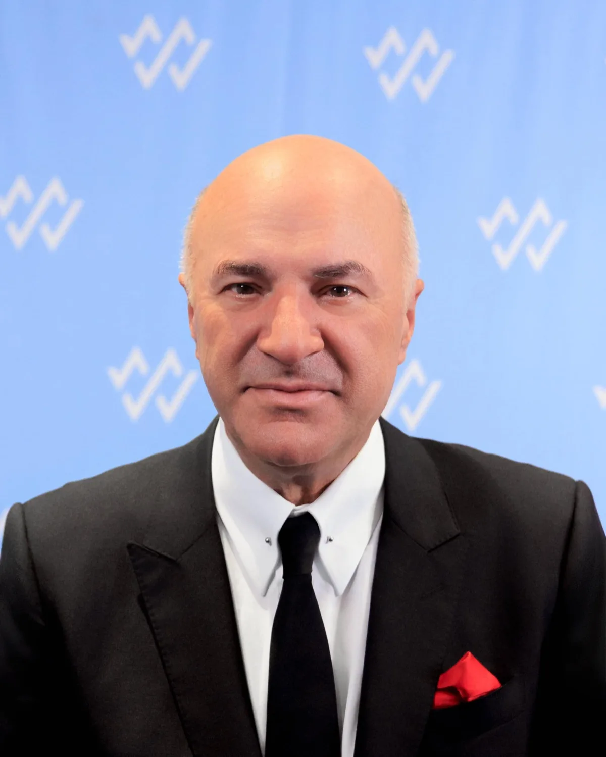 Bald man in black suit, white shirt and red pocket square against light blue backdrop