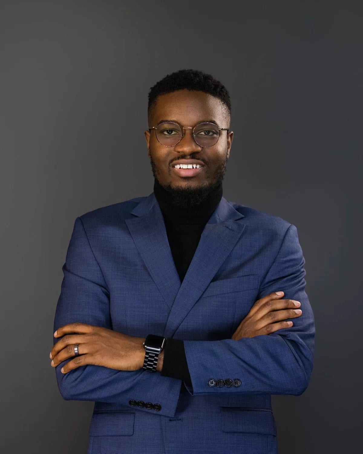 Smiling man in blue blazer, black turtleneck and round glasses, arms crossed against grey backdrop