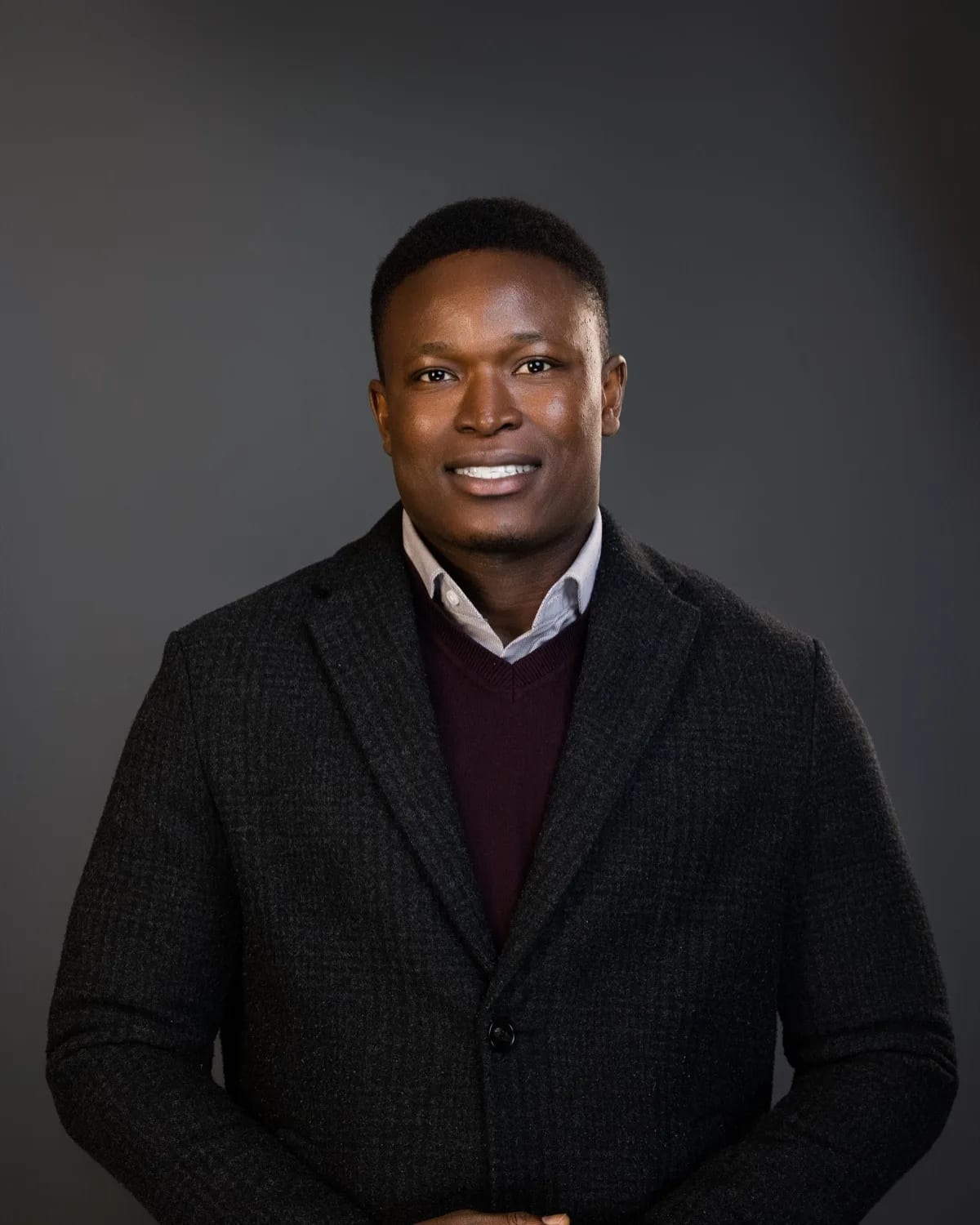 Smiling man in charcoal tweed blazer, burgundy sweater and light shirt against grey studio backdrop