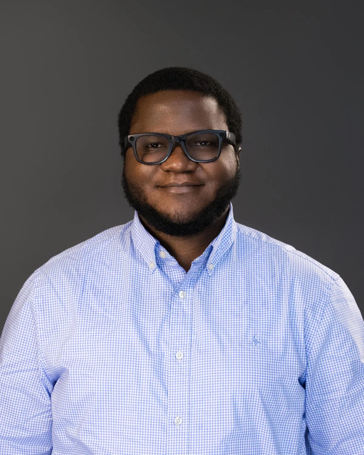 Bearded man in glasses and light blue checked shirt smiling against dark grey studio backdrop