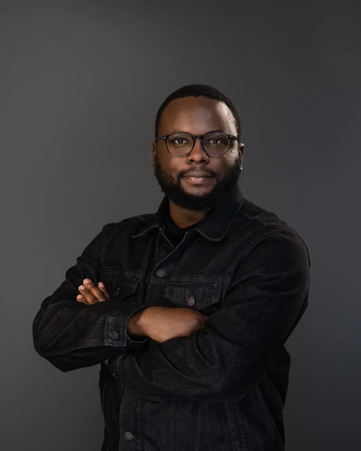 Man with glasses and beard in black denim jacket, arms crossed, against grey studio backdrop