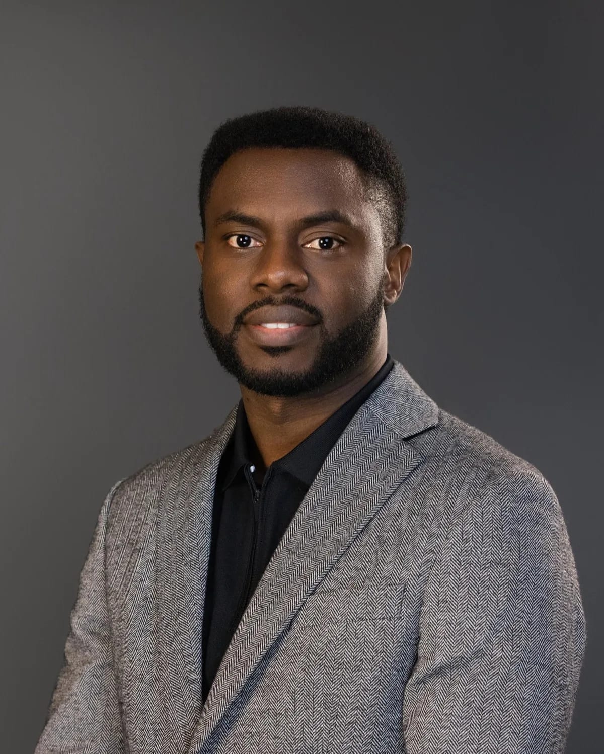 Man with beard in gray herringbone blazer over black shirt against dark gray studio backdrop