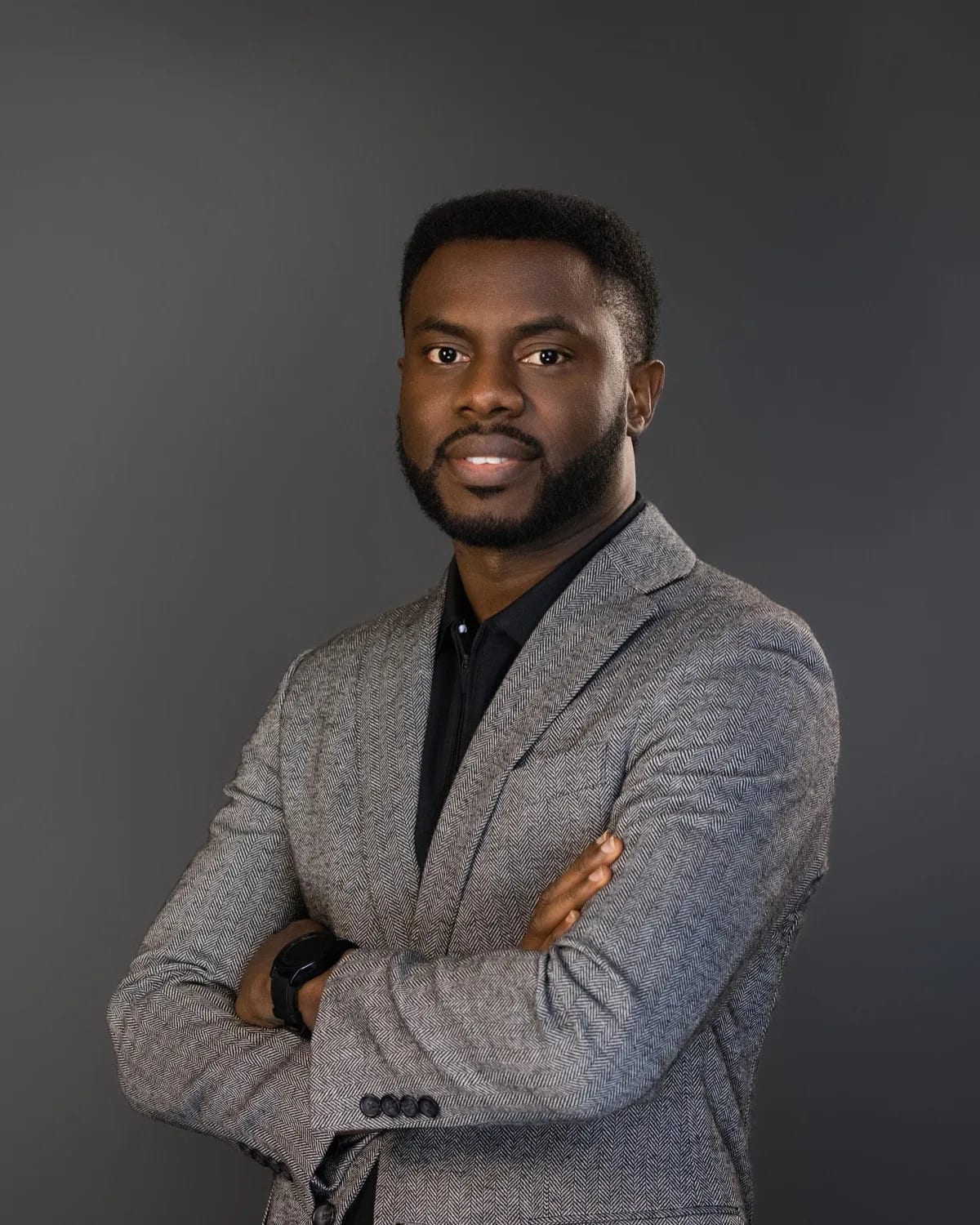 Bearded man in grey herringbone blazer over black shirt, arms crossed against dark grey backdrop