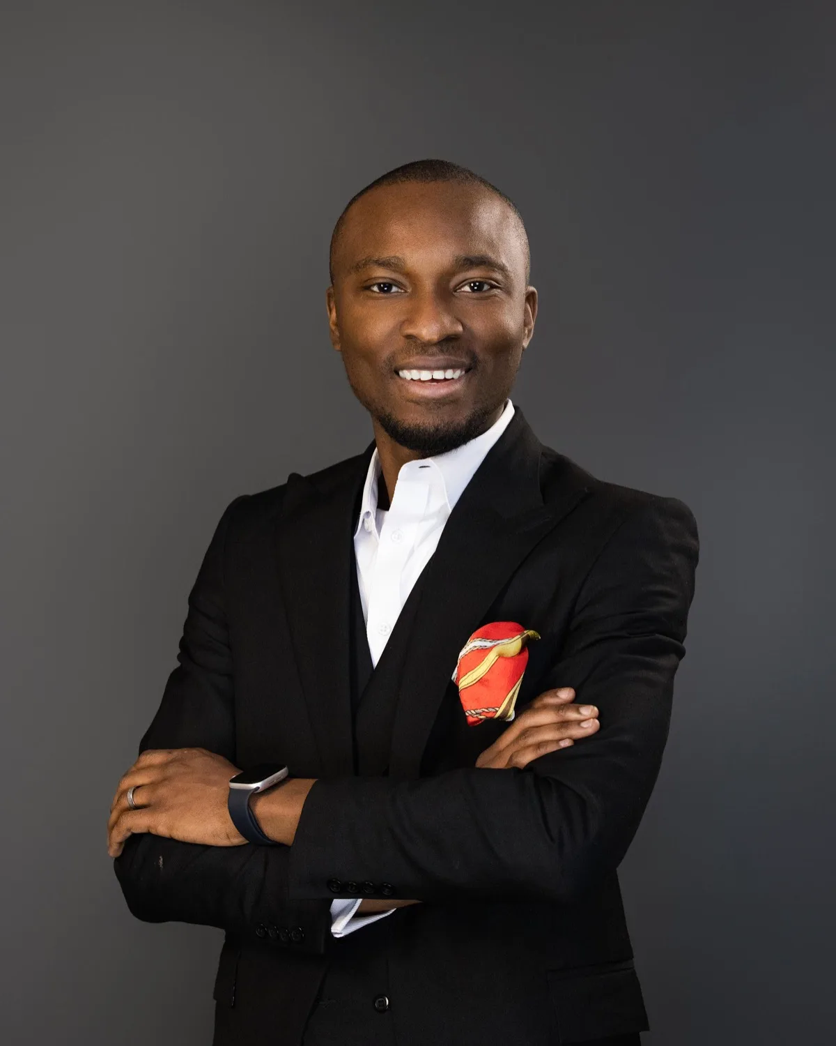 Smiling man in black suit with white shirt and red pocket square against gray backdrop
