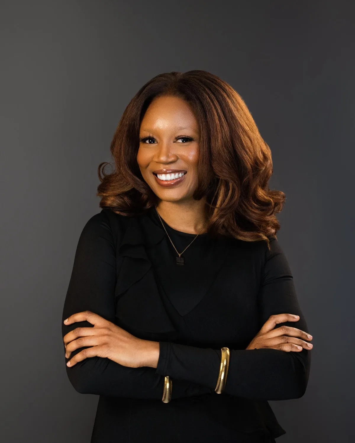 Smiling woman with auburn wavy hair and gold bangles, arms crossed, against dark grey backdrop