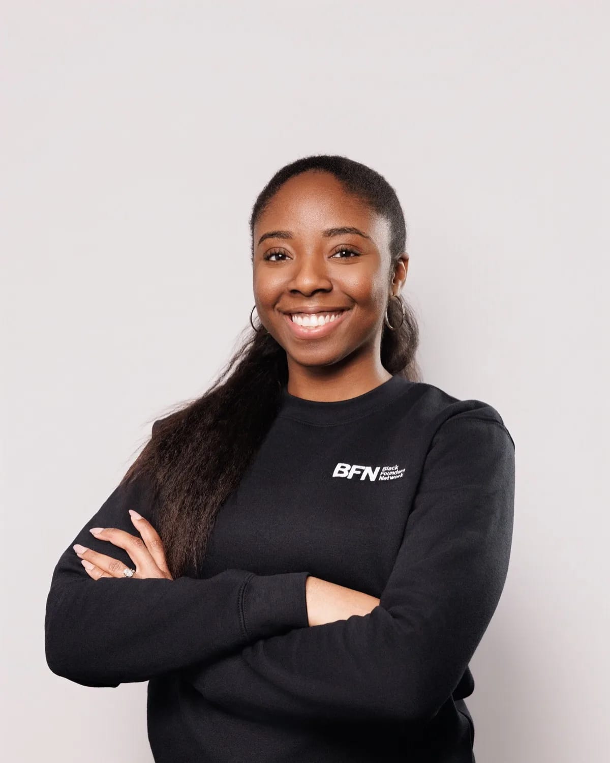 Smiling woman with long hair and hoop earrings, arms crossed, in black sweatshirt against gray backdrop
