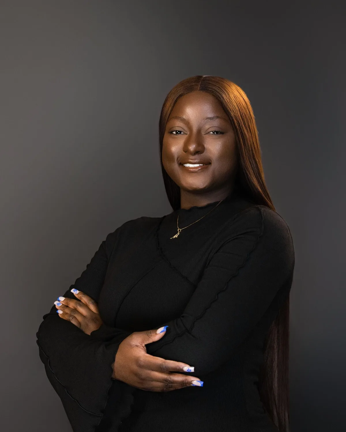 Smiling woman with long auburn hair in black top, arms crossed, against grey studio backdrop