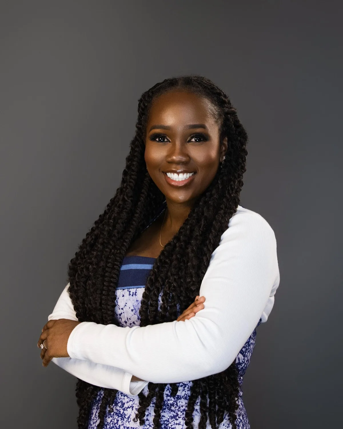 Smiling woman with long twist braids and arms crossed, white top over blue patterned dress