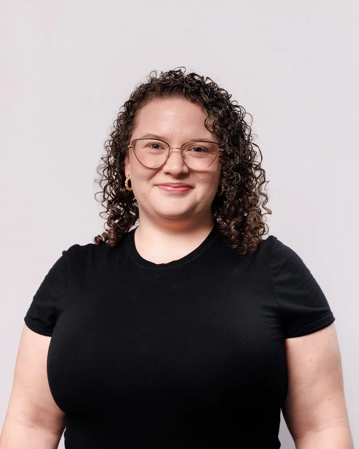 Woman with curly hair and gold cat-eye glasses in black t-shirt against light grey backdrop