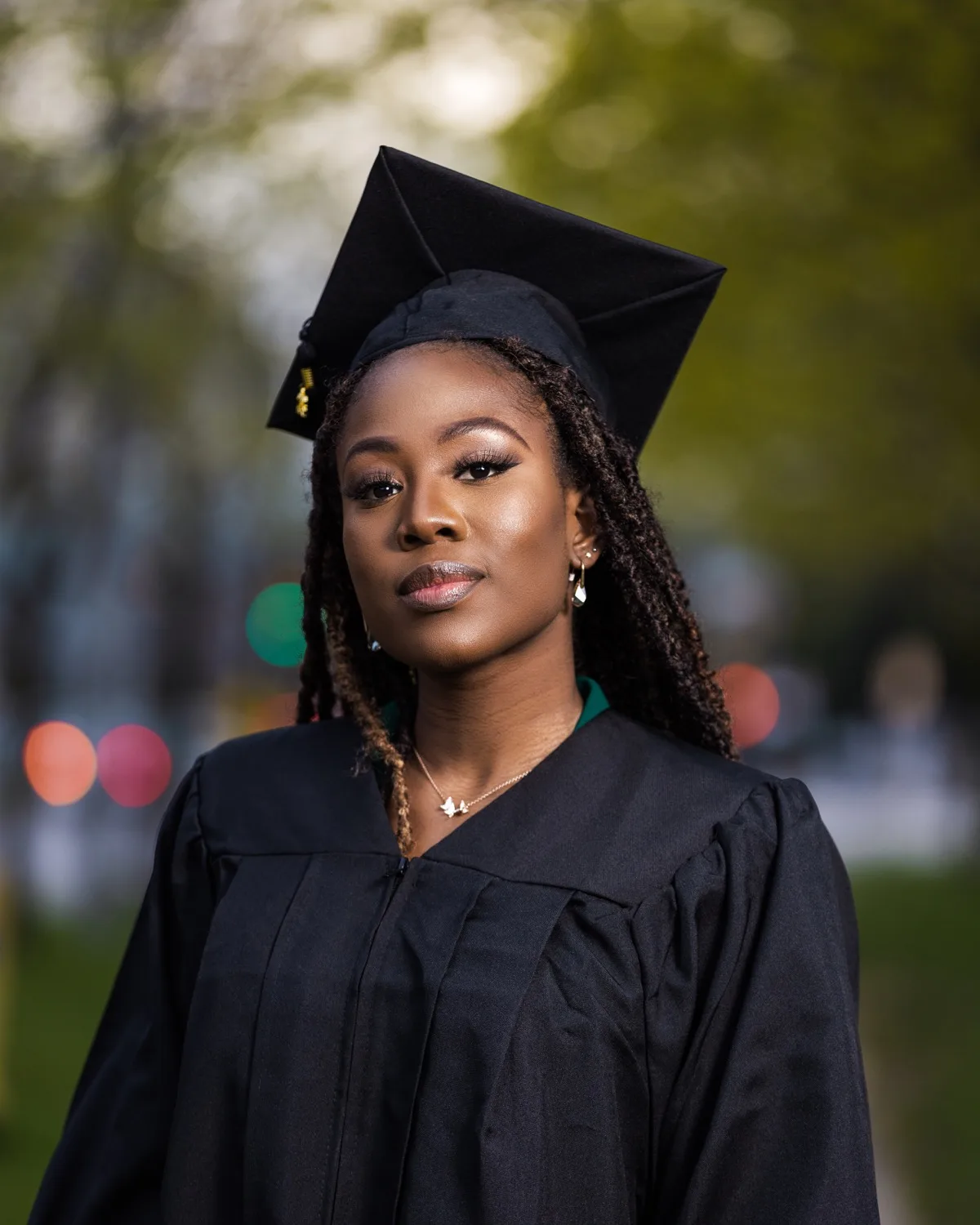 Young woman in black graduation cap and gown outdoors with softly blurred green foliage behind